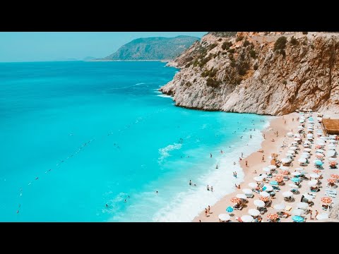 Aerial view of a crowded beach along The Lycian Way with turquoise water by a rugged coastline, featuring sunbathers under umbrellas and cliffs on one side.